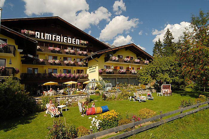 Ein großes Alpenhotel mit blumengeschmückten Balkonen, einem grünen Rasen mit Gartenmöbeln und Menschen, die sich an einem sonnigen Tag im Freien entspannen.