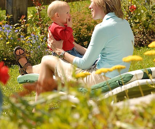 Ein Erwachsener und ein Baby sitzen zusammen auf einem Liegestuhl in einem Garten, einander zugewandt und lächelnd, mit Blumen und einem Holzzaun im Hintergrund.