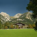 Auf einer grünen Wiese, umgeben von Bäumen und vor der Kulisse schroffer, bewaldeter Berge, steht ein Gebäude im Stil einer Lodge unter einem strahlend blauen Himmel.