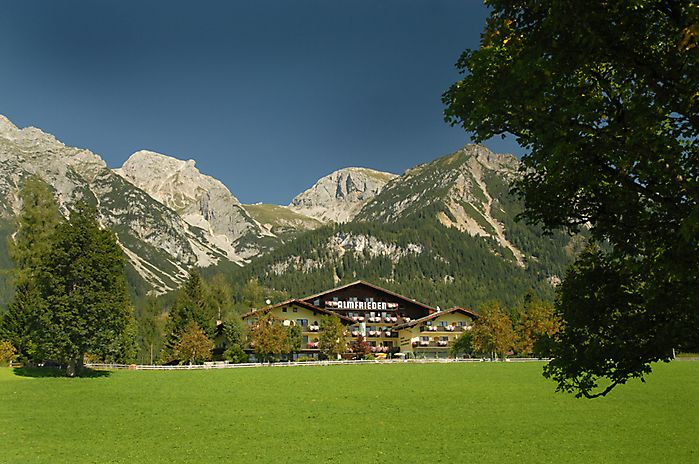 Auf einer grünen Wiese, umgeben von Bäumen und vor der Kulisse schroffer, bewaldeter Berge, steht ein Gebäude im Stil einer Lodge unter einem strahlend blauen Himmel.