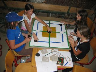 Vier Mädchen sitzen um einen Tisch und spielen ein Brettspiel mit Karten und runden Spielsteinen auf einem selbstgemachten Spielbrett.