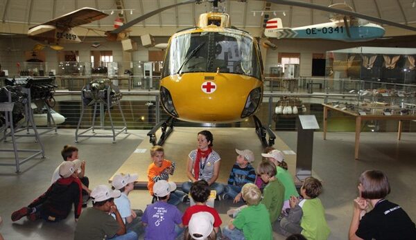 Eine Gruppe Kinder sitzt vor einem gelben Rettungshubschrauber in einem Museum und hört einer Frau zu.
