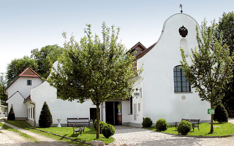 Ein weißes, historisches Gebäude mit Rundbogenfenstern und einem geschwungenen Giebel, umgeben von Bäumen, Bänken und einem Kopfsteinpflasterweg, der zum Eingang führt.