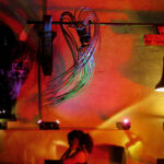 Eine Person steht in einem Innenraum unter bunten Lichtern, über ihr sind abstrakte Wandkunst, eine Glocke und eine Maske oder Skulptur zu sehen.