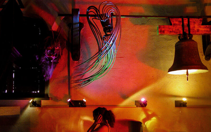 Eine Person steht in einem Innenraum unter bunten Lichtern, über ihr sind abstrakte Wandkunst, eine Glocke und eine Maske oder Skulptur zu sehen.