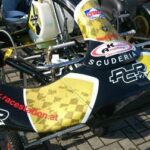 Nahaufnahme der Frontpartie eines gelb-schwarzen Go-Karts mit den Aufschriften "SCUDERIA" und "ACR", das auf einer gepflasterten Fläche abgestellt ist.
