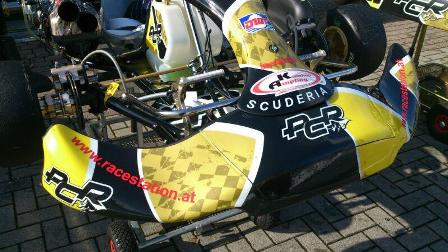 Nahaufnahme der Frontpartie eines gelb-schwarzen Go-Karts mit den Aufschriften "SCUDERIA" und "ACR", das auf einer gepflasterten Fläche abgestellt ist.