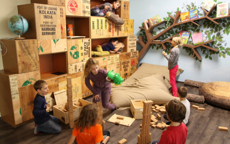 Kinder spielen mit Holzklötzen und Kisten in einem Raum mit einem baumförmigen Bücherregal, Büchern und Kissen. Einige Kinder bauen, andere erkunden die Umgebung oder sitzen auf einem großen Sitzsack.
