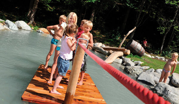 Fünf Kinder stehen auf einem Holzfloß in einem flachen Teich und halten sich an einem roten Seil fest, während ein anderes Kind auf Felsen am Wasser steht.