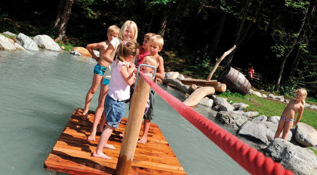 Fünf Kinder stehen auf einem Holzfloß in einem flachen Teich und halten sich an einem roten Seil fest, während ein anderes Kind auf Felsen am Wasser steht.
