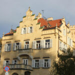 Ein beiges, vierstöckiges historisches Gebäude mit weißen Fensterläden, dekorativem Balkon und einem roten Ziegeldach, fotografiert bei Tageslicht mit einigen Bäumen und einem Parkverbotsschild davor.