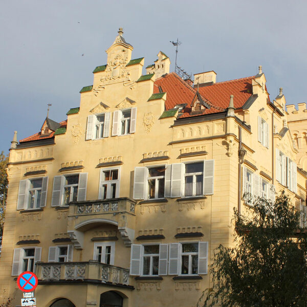 Ein beiges, vierstöckiges historisches Gebäude mit weißen Fensterläden, dekorativem Balkon und einem roten Ziegeldach, fotografiert bei Tageslicht mit einigen Bäumen und einem Parkverbotsschild davor.