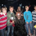 Eine Gruppe von fünf Jungen steht in einem Raum mit Sternenhimmel im Hintergrund um eine Person im Kostüm eines gehörnten Monsters herum; ein Junge macht ein Peace-Zeichen.