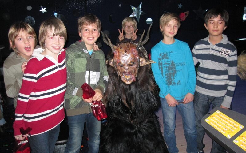 Eine Gruppe von fünf Jungen steht in einem Raum mit Sternenhimmel im Hintergrund um eine Person im Kostüm eines gehörnten Monsters herum; ein Junge macht ein Peace-Zeichen.