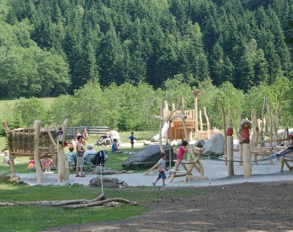 Kinder und Erwachsene befinden sich auf einem Außenspielplatz mit Holzkonstruktionen, umgeben von Gras und Bäumen, mit einem bewaldeten Hügel im Hintergrund.