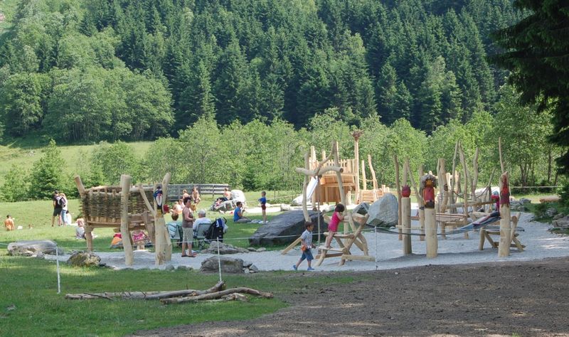 Kinder und Erwachsene befinden sich auf einem Außenspielplatz mit Holzkonstruktionen, umgeben von Gras und Bäumen, mit einem bewaldeten Hügel im Hintergrund.