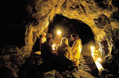 Vier Personen sitzen auf Bänken in einer schwach beleuchteten Höhle oder Mine, die von mehreren um sie herum aufgestellten Laternen beleuchtet wird.