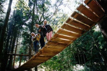 Drei Personen stehen auf einer schmalen hölzernen Hängebrücke in einem Waldgebiet, umgeben von hohen Bäumen und Grün.
