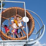 Vier Kinder stehen in einem großen, kreisrunden Seilklettergerüst und lächeln an einem sonnigen Tag mit blauem Himmel im Hintergrund.