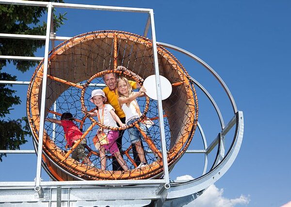 Vier Kinder stehen in einem großen, kreisrunden Seilklettergerüst und lächeln an einem sonnigen Tag mit blauem Himmel im Hintergrund.