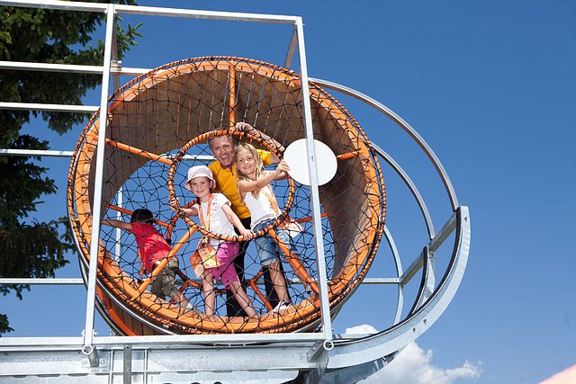 Vier Kinder stehen in einem großen, kreisrunden Seilklettergerüst und lächeln an einem sonnigen Tag mit blauem Himmel im Hintergrund.