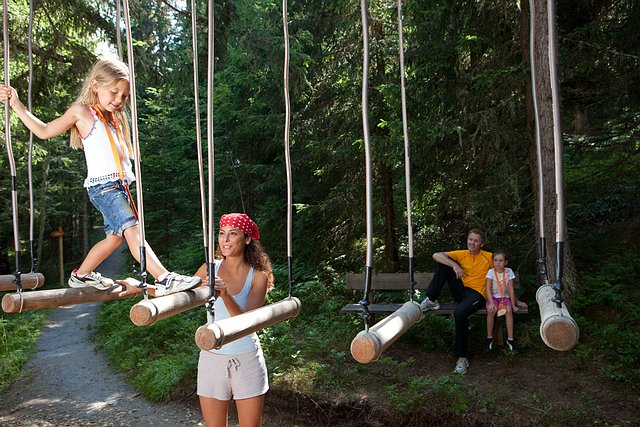 Eine Frau hilft einem Kind beim Überqueren einer Baumstammschaukel in einem Wald, während ein anderer Erwachsener und ein Kind von einer Bank im Hintergrund zusehen.