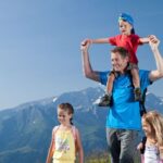 Ein Mann trägt ein Kind auf seinen Schultern und geht mit zwei anderen Kindern auf einem Bergpfad in der Nähe eines gelben Wegweisers, im Hintergrund sind schneebedeckte Berge zu sehen.