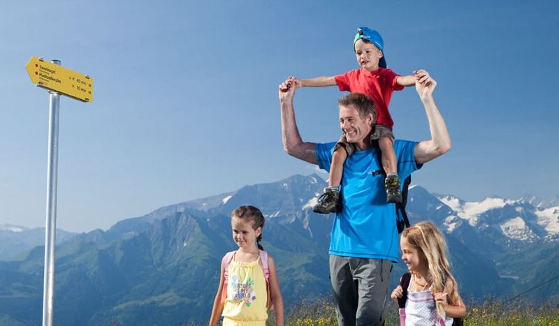 Ein Mann trägt ein Kind auf seinen Schultern und geht mit zwei anderen Kindern auf einem Bergpfad in der Nähe eines gelben Wegweisers, im Hintergrund sind schneebedeckte Berge zu sehen.