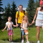 Eine vierköpfige Familie, zwei Erwachsene und zwei Kinder, geht an einem sonnigen Tag gemeinsam auf einem Graspfad in einem Waldgebiet spazieren.