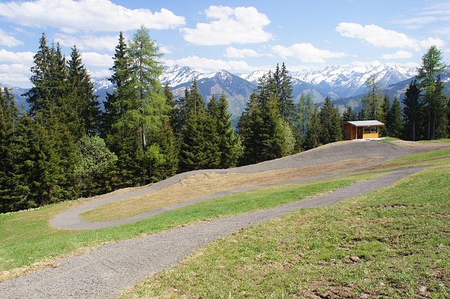Ein gewundener Schotterweg führt durch eine Grasfläche mit Kiefern, einer kleinen Holzhütte und schneebedeckten Bergen im Hintergrund unter einem teilweise bewölkten Himmel.