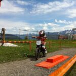 Ein Kind mit Helm fährt mit einem kleinen Dirtbike auf einer eingezäunten Strecke im Freien, im Hintergrund sind Berge und ein See zu sehen.