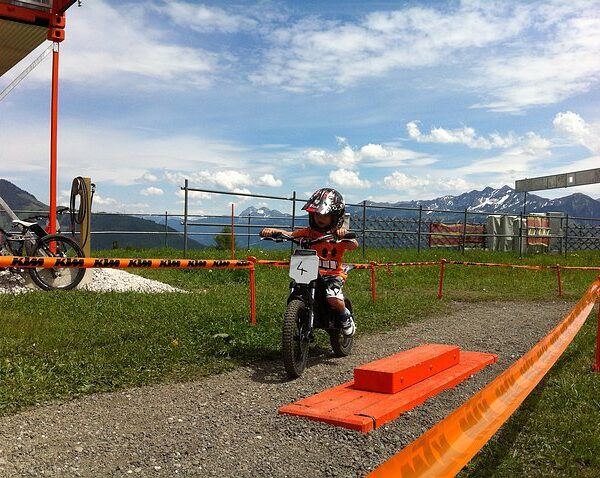 Ein Kind mit Helm fährt mit einem kleinen Dirtbike auf einer eingezäunten Strecke im Freien, im Hintergrund sind Berge und ein See zu sehen.