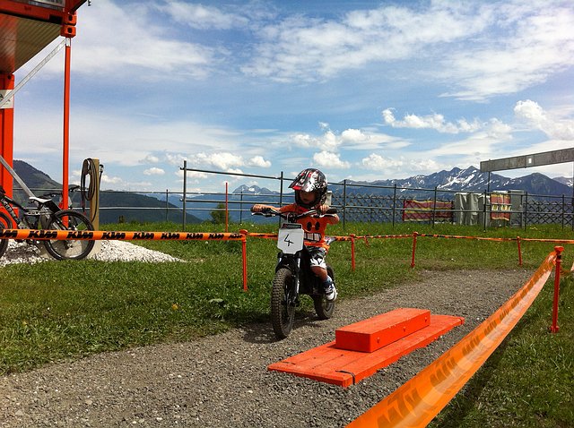 Ein Kind mit Helm fährt mit einem kleinen Dirtbike auf einer eingezäunten Strecke im Freien, im Hintergrund sind Berge und ein See zu sehen.