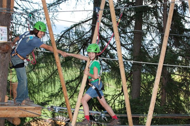 Zwei Personen mit Helmen und Gurten geben sich auf einem Hochseilgarten zwischen Bäumen die Hand, an dessen Struktur Sicherheitsleinen befestigt sind.