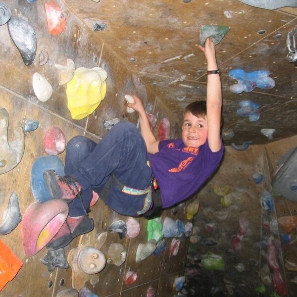 Ein Kind in einem lila T-Shirt klettert eine Indoor-Boulderwand hoch, klammert sich an bunte Griffe und schaut in die Kamera.