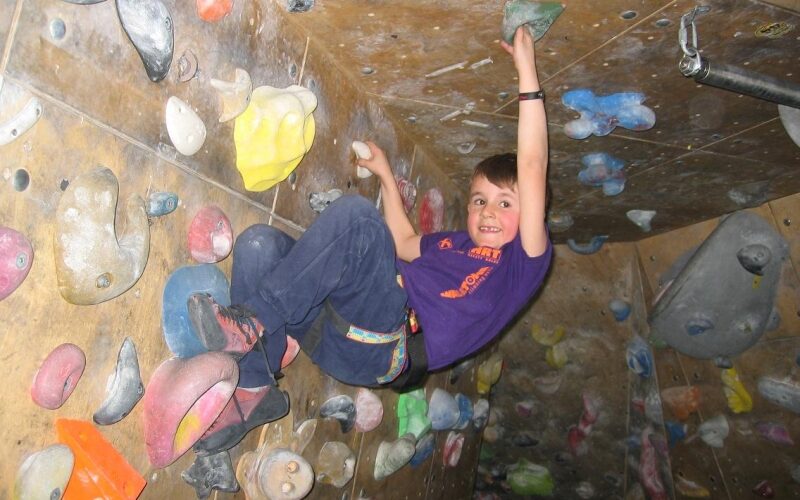 Ein Kind in einem lila T-Shirt klettert eine Indoor-Boulderwand hoch, klammert sich an bunte Griffe und schaut in die Kamera.