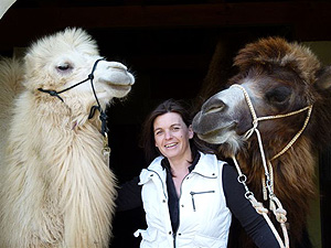 Eine Frau steht lächelnd zwischen einem weißen Kamel zu ihrer Linken und einem braunen Kamel zu ihrer Rechten, beide tragen Halfter.