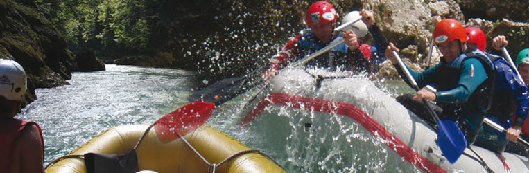 Eine Gruppe von Menschen mit Helmen und Schwimmwesten paddelt auf einem Wildwasserfloß durch einen Fluss und spritzt dabei Wasser auf, während sie durch die Stromschnellen navigieren.
