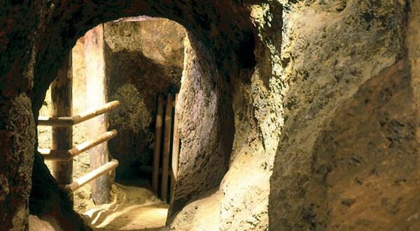 Ein schmaler, in den Fels gehauener Tunnel mit auf der linken Seite sichtbaren Holzgeländern, die mit künstlicher Beleuchtung beleuchtet werden.