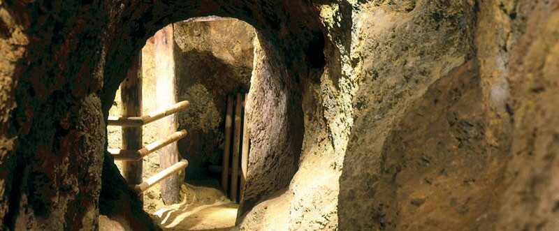 Ein schmaler, in den Fels gehauener Tunnel mit auf der linken Seite sichtbaren Holzgeländern, die mit künstlicher Beleuchtung beleuchtet werden.