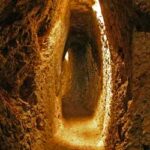 Schmaler, schwach beleuchteter, in den Fels gehauener Tunnel mit rauen, unebenen Wänden und einem Erdboden, beleuchtet durch Licht aus einer unsichtbaren Quelle.