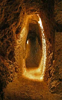 Schmaler, schwach beleuchteter, in den Fels gehauener Tunnel mit rauen, unebenen Wänden und einem Erdboden, beleuchtet durch Licht aus einer unsichtbaren Quelle.