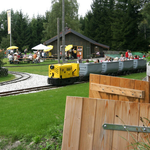 Ein kleiner gelber Zug mit offenen Personenwagen fährt auf den Gleisen in der Nähe eines Holzgebäudes mit Sitzgelegenheiten im Freien, umgeben von Gras und Bäumen.