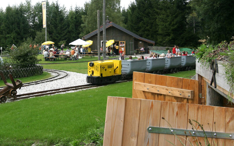 Ein kleiner gelber Zug mit offenen Personenwagen fährt auf den Gleisen in der Nähe eines Holzgebäudes mit Sitzgelegenheiten im Freien, umgeben von Gras und Bäumen.