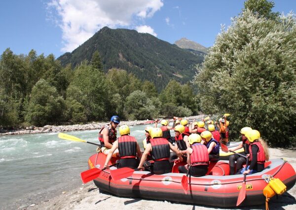 Eine Gruppe von Menschen mit Helmen und Schwimmwesten sitzt in einem roten Schlauchboot an einem Fluss und bereitet sich mit einem Führer auf eine Wildwasserfahrt vor; im Hintergrund sind Berge und Bäume zu sehen.