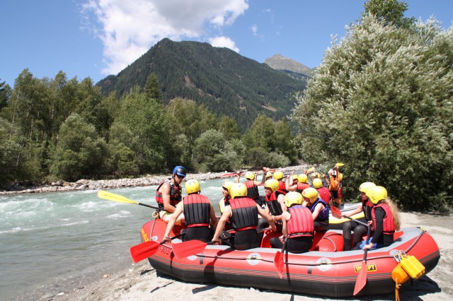 Eine Gruppe von Menschen mit Helmen und Schwimmwesten sitzt in einem roten Schlauchboot an einem Fluss und bereitet sich mit einem Führer auf eine Wildwasserfahrt vor; im Hintergrund sind Berge und Bäume zu sehen.