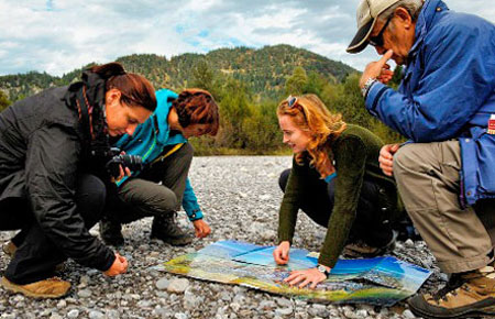 Vier Personen knien im Freien auf felsigem Boden, betrachten eine große, vor ihnen ausgebreitete Karte und zeigen darauf, im Hintergrund sind Bäume und Hügel zu sehen.