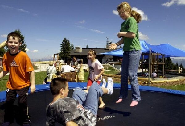 Vier Kinder spielen und springen im Freien auf einem Trampolin, im Hintergrund sind Erwachsene und Spielgeräte zu sehen.