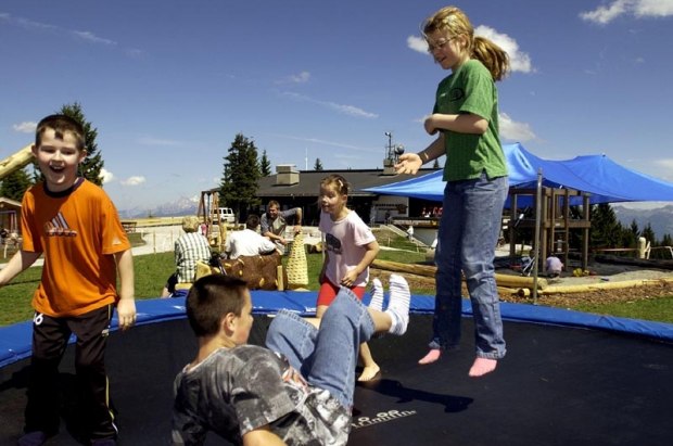 Vier Kinder spielen und springen im Freien auf einem Trampolin, im Hintergrund sind Erwachsene und Spielgeräte zu sehen.
