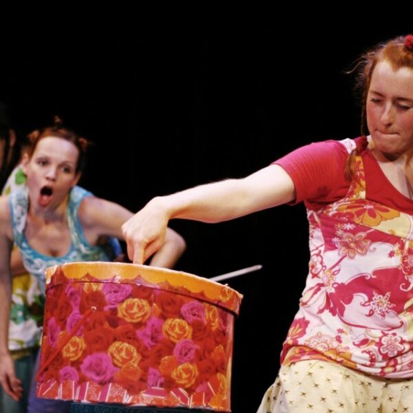 Drei Frauen auf der Bühne; eine vorne steckt ihre Hand in eine verzierte Trommel, während zwei andere im Hintergrund mit überraschten Gesichtern zuschauen.